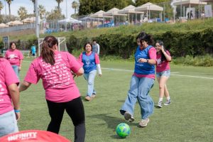 Girls wearing pink Girls Empowerment Day shirts play soccer during a youth sports clinic on an outdoor field.