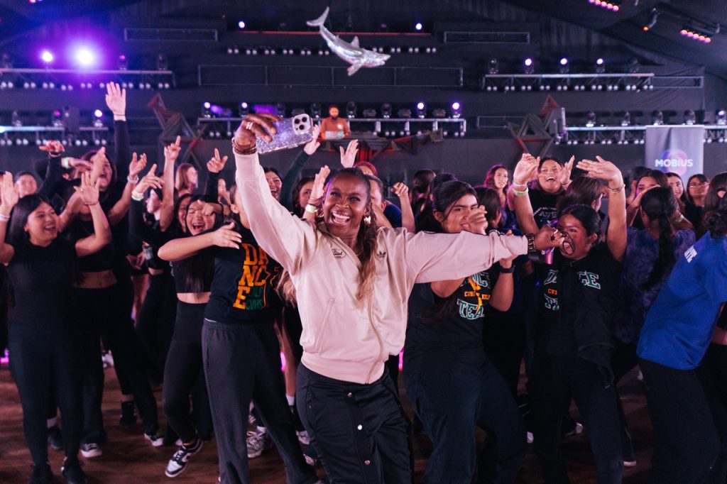 A person in a light jacket holds up a phone for a selfie while surrounded by a large group dancing with raised arms inside a concert-style venue with stage lighting overhead.