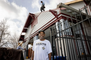 A person in a New Orleans Saints t-shirt standing in front of a house with people doing work on it