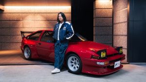 Sung Kang leaning against a red sports car with pop-up headlights in a modern garage