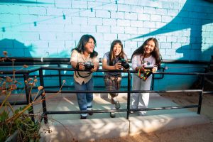 Three young people hold cameras and stand together along a railing in front of a bright blue brick wall.